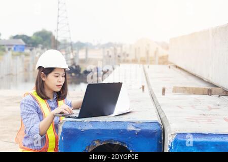 Beautiful Asian female engineer in white safety hard hat using laptop computer notebook doing job at construction site outside office. Idea for modern Stock Photo