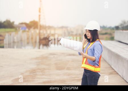 Beautiful Asian female engineer in white safety hard hat doing job at construction site outside office. Idea for modern working woman hightway road Stock Photo
