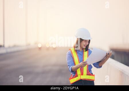 Beautiful Asian female engineer in white safety hard hat doing job at construction site outside office. Idea for modern working woman hightway road Stock Photo