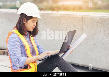 Beautiful Asian female engineer in white safety hard hat using laptop computer notebook doing job at construction site outside office. Idea for modern Stock Photo