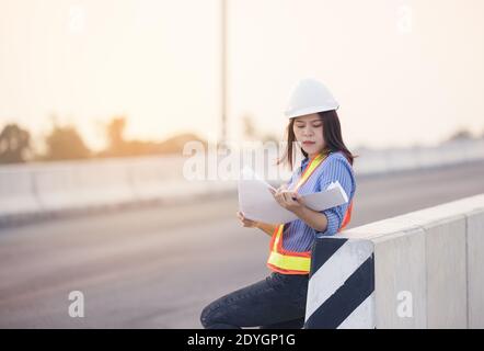 Beautiful Asian female engineer in white safety hard hat doing job at construction site outside office. Idea for modern working woman hightway road Stock Photo