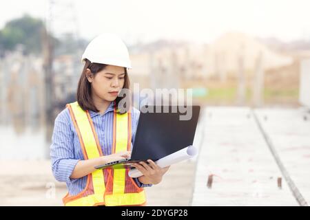 Beautiful Asian female engineer in white safety hard hat using laptop computer notebook doing job at construction site outside office. Idea for modern Stock Photo