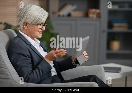 Senior businesswoman in eyeglasses sitting on sofa and talking online with partner using digital tablet Stock Photo