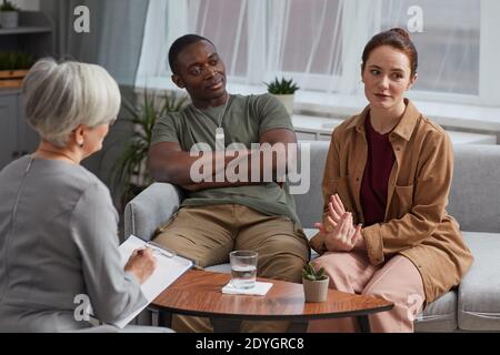 Young woman talking about her problems to psychologist while sitting on sofa together with husband during consultation Stock Photo