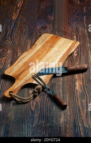 Shabby wooden table with empty cutting board and old forged knife and fork for barbecue Stock Photo