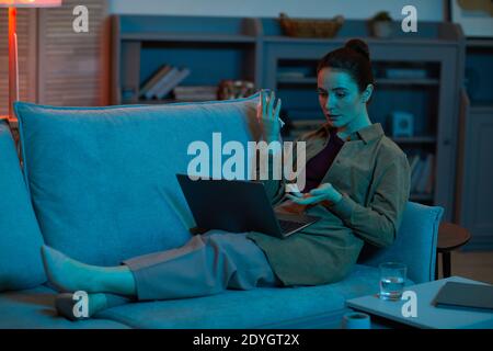 Young woman lying on sofa with laptop on her legs and talking online with her friend in the room at home Stock Photo