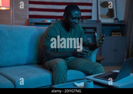 African young man drinking alcohol and has a lively conversation online using his laptop at home Stock Photo