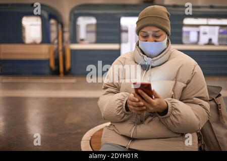Young woman in mask and in warm clothing sitting in subway and using her mobile phone while waiting for her train Stock Photo