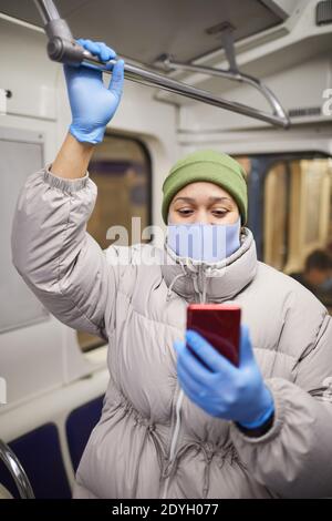 Young woman in protective gloves and mask using mobile phone while standing on a train Stock Photo