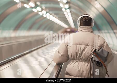 Rear view of plump woman in headphones with bag behind her back riding on escalator Stock Photo
