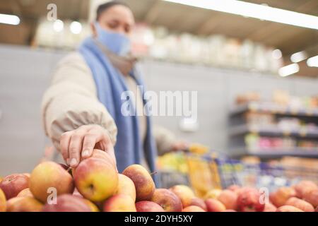 Young woman in mask choosing fresh apples while doing shopping in supermarket Stock Photo