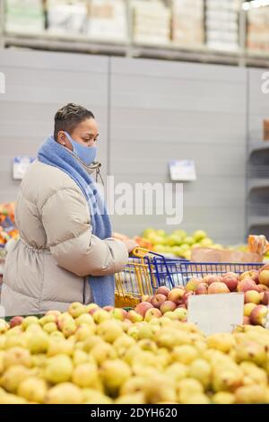 Young woman in warm clothing and in mask buying fresh fruits in supermarket Stock Photo