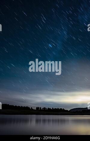 night landscape with a sky with stars trail, with the silhouette of a forest in the background and a lake in the foreground, vertical Stock Photo