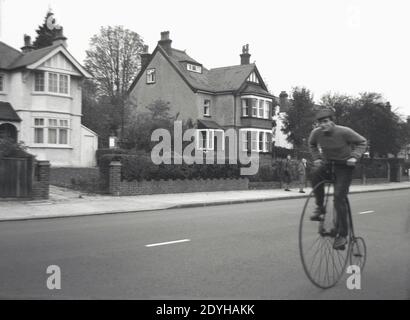 1960s, historical, a man riding an old, early type of bicycle, a 'penny-farthing' or 'ordinary' on a suburban road, England, UK. Also known as a high wheel or high wheeler, the bicycle with its large front wheel was popular in the 1870s and early 80s,  providing speed and comfort. Although dangerous to ride because of its height, the bike became a symbol of the late victorian era and the birth of cycling as a sport. It became obsolete in the late 1980s, with the creation of the modern bicycle with the invention of chain-driven gear trains and pneumatic tires. Stock Photo