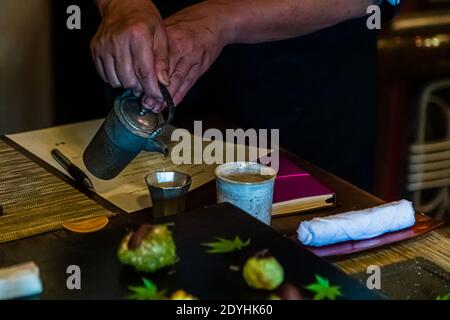 Rakan, a kaiseki restaurant run by chef Atsuko Kato, on the Izu Peninsula, Japan. A hand is pouring a drink from a small metal jug into a cup; next to it lie a menu and a notebook Stock Photo