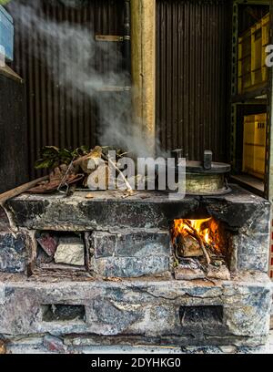 Rakan, a kaiseki restaurant run by chef Atsuko Kato, on the Izu Peninsula, Japan. Rice simmers in a metal pot on a traditional wood-fired stone stove, smoke rising. Stock Photo