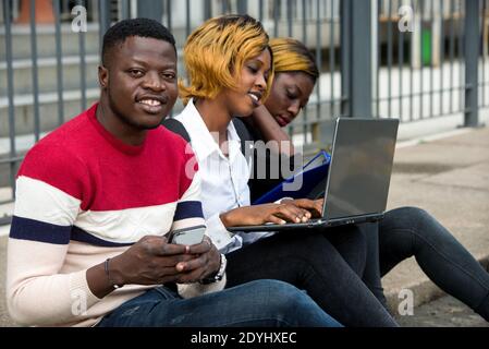 Group of attractive smiling young casual dressed students using laptop sitting outdoors on campus of university. Stock Photo