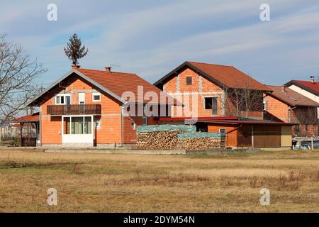 Row of newly built unfinished red brick family houses with new roof tiles surrounded with dry grass and small wooden outdoor storage sheds on cloudy Stock Photo