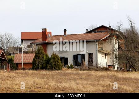White suburban family house damaged by shrapnel during war with dilapidated facade and repaired roof with new roof tiles on top of small hill Stock Photo