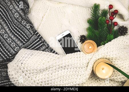 Modern winter cozy background, woolen and knitted sweater, fir branch with red berries and candles, blank smartphone Stock Photo