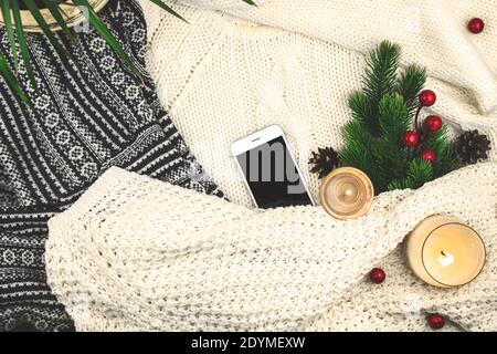 Modern winter cozy background, woolen and knitted sweater, fir branch with red berries and candles, blank smartphone Stock Photo