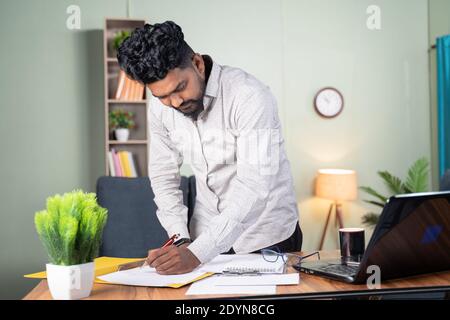 Young business man busy in paperwork by referring documents and laptop - concept of preparing plan, managing tasks by standing at office table. Stock Photo