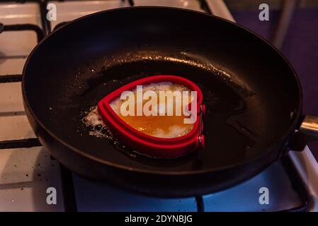 pan-fried egg in the shape of a heart on a white gas stove and black grill, which holds the pan over a fire flame Stock Photo
