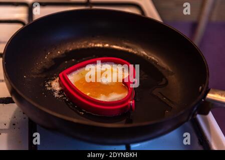 pan-fried egg in the shape of a heart on a white gas stove and black grill, which holds the pan over a fire flame Stock Photo