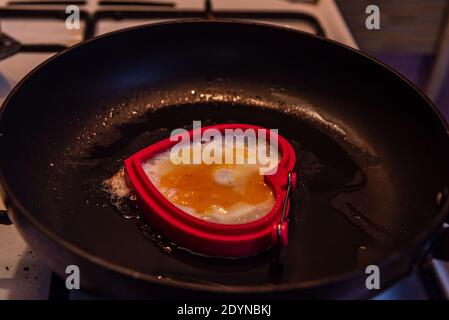 pan-fried egg in the shape of a heart on a white gas stove and black grill, which holds the pan over a fire flame Stock Photo