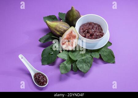 Fig jam with figs, fig leaves and white spoon with jam on purple background. Stock Photo