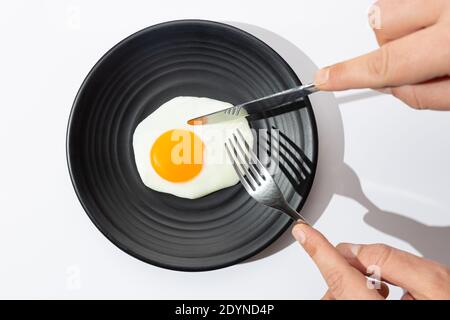 Hands are going to eat with knife and fork a fried egg on black plate on  white background. Stock Photo