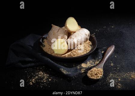 Pieces of ginger and ginger powder, in  wooden spoon and plate,  on black background. Stock Photo