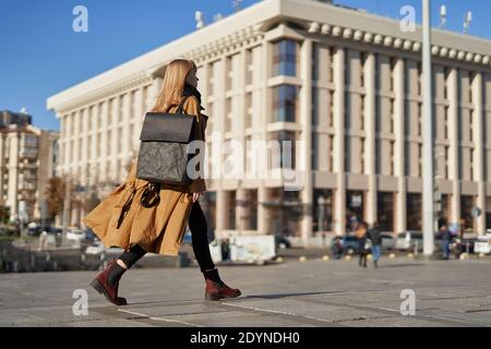 Blonde girl in orange trench walking with black ecological paper backpack in European city Stock Photo