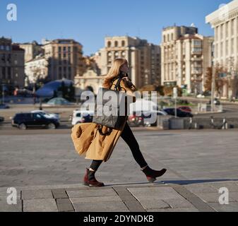 Attractive blonde woman in orange trench walking in a hurry with recycled made paper backpack on blue sky background of the european city streets. Bus Stock Photo