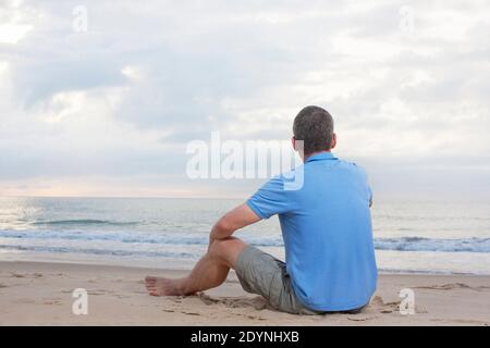 Mature man in blue t-shirt sitting on a beach looking at the sea at sunrise Stock Photo