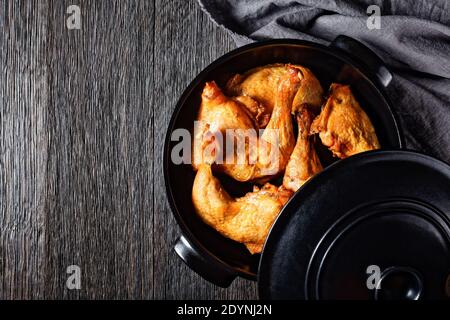 crispy roasted chicken legs in black ceramic pan on a dark wooden table, horizontal view from above, flat lay, free space Stock Photo