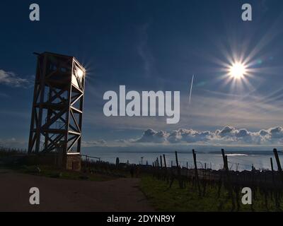 Wooden observation tower on vineyard slopes above small village Hagnau am Bodensee, Lake Constance, Germany with bright sun in backlit and reflection. Stock Photo