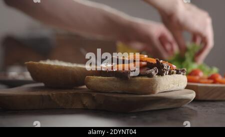 Man put steak slices on on small ciabatta bun for sandwich on kitchen countertop, wide photo Stock Photo