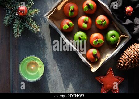 Marzipan sponge apples. Tasty Christmas dessert with fir twigs, green candle, pine cone, soft star. Top view on in rustic wooden tray on textile cotto Stock Photo