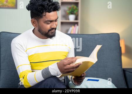 concentrated young man reading book during leisure time at home by sitting on sofa - concept of reading hobby. Stock Photo