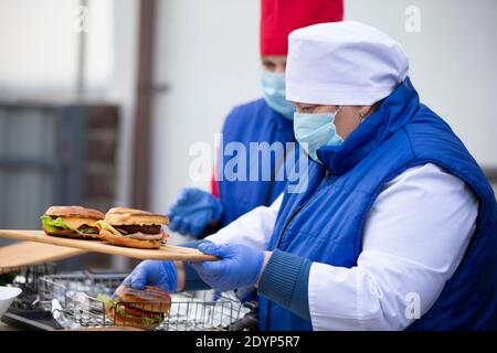 Cooking street food during the coronavirus panademy. A chef in a medical mask prepares hamburgers. Stock Photo