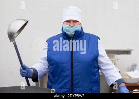 November 13, 2020. Belarus, Gomil region. PA Cooking street food during the coronavirus panademy. A chef in a medical mask with a large scoop. Stock Photo