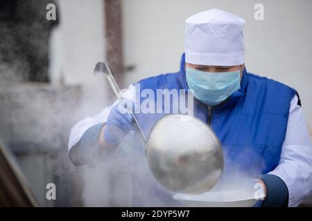 A chef in a medical mask with a large scoop pours Stock Photo
