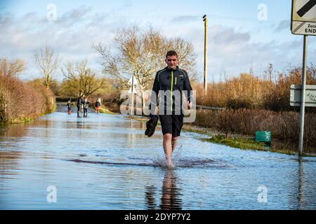 Wytham, Oxfordshire, UK. 27 December, 2020. Euan has to remove his shoes to traverse the flooded road on his run to Wytham. Flooding in Oxfordshire. Storm Bella brought even more rain to Oxfordshire causing flooding in low lying areas. Plenty of people are out exercising in the sunshine. Credit: Sidney Bruere/Alamy Live News Stock Photo