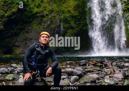 A closeup portrait of a cheerful man rock climber in safety harness sitting on a rock with a waterfall behind Stock Photo