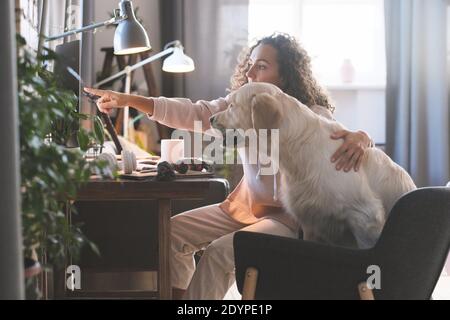 Young woman sitting with her dog in front of computer and pointing at computer showing something to it Stock Photo
