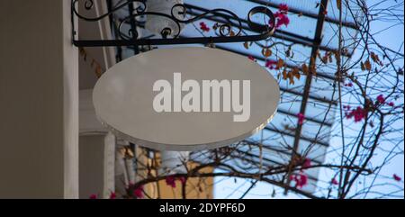 Blank white metal oval wall sign, commercial mockup, template. Empty billboard cutout hanging from black metal base outside of store, copyspace. Blur Stock Photo