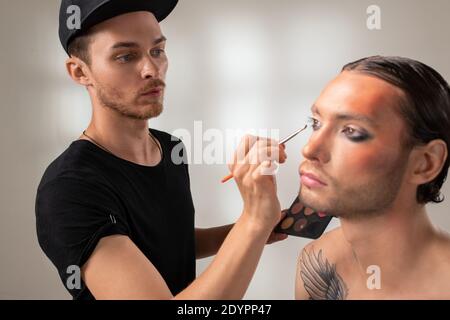 Young makeup artist with brush or applicator applying grey shadow on eyelids of male fashion model with tattoo on shoulder in studio Stock Photo
