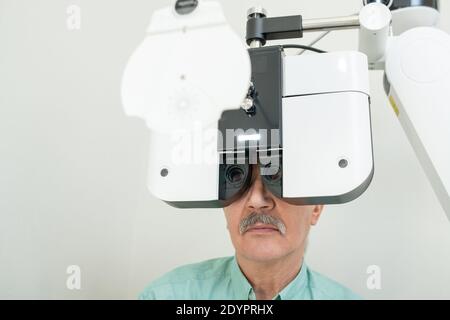 Senior male patient sitting in front of ophthalmological equipment and looking through lens during medical check-up of his eyesight in clinics Stock Photo