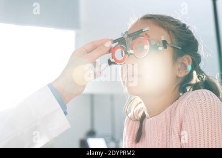 Elementary schoolgirl sitting in front of ophthalmologist during check-up of her eyesight with optometrist trial frame in contemporary clinics Stock Photo
